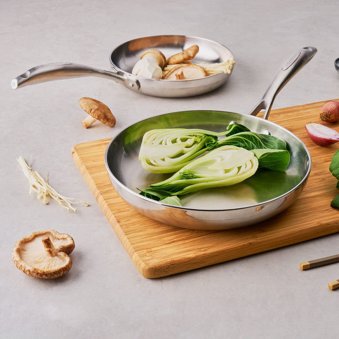 Stainless steel frying pan with bok choy on a wooden cutting board, surrounded by ingredients on a gray surface.
