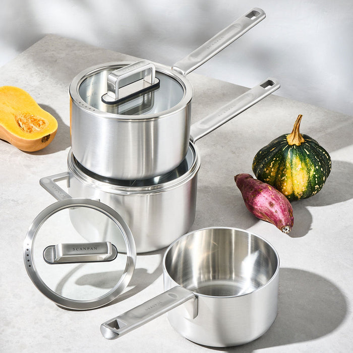 Set of stainless steel cookware on a light surface with vegetables.
